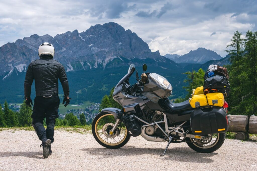 Motorrad mit voll bepacktem Tankrucksack wasserdicht vor Alpenkulisse, Fahrer geht Richtung Aussichtspunkt